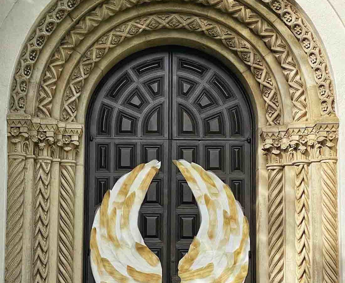 Ornate door. 3 golden arches surround a black carved wooden door. Upon the door are two golden angel wings.