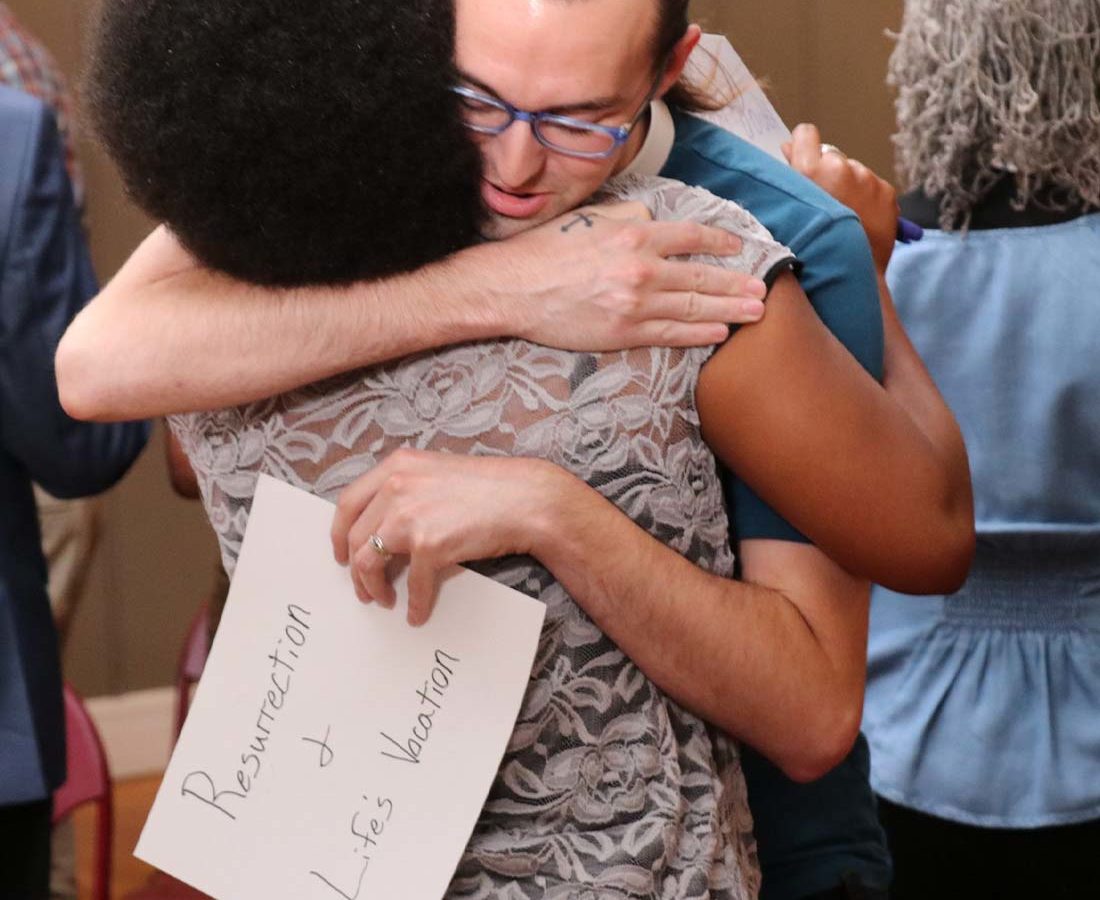 A woman of color in a patterned blouse hugs a white man in a blue shirt holding a piece of paper.