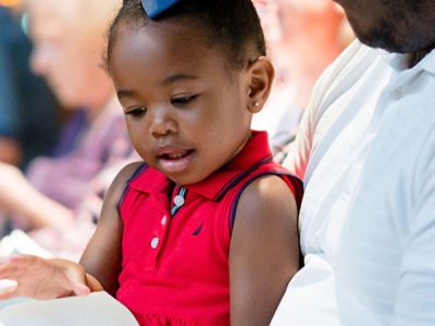 A father holds his young child wearing a red shirt on his lap. Together they are looking at a service bulletin.