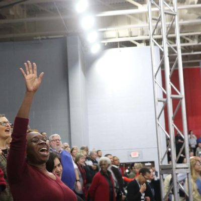 People Worshiping. A woman of color in a red shirt is singing with one arm raised overhead in the foreground.