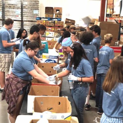 Several college aged students wearing blue shirts are packing care packages in an assembly line.