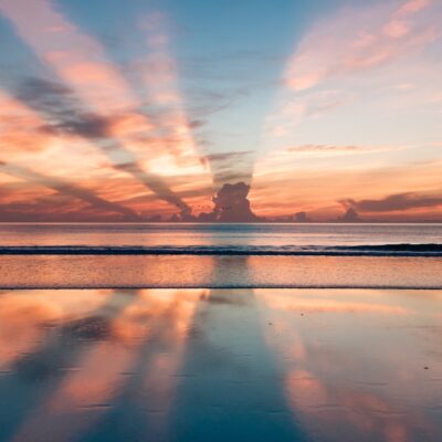Sun beams streak across the sky as small waves lap upon the shore of a beach.