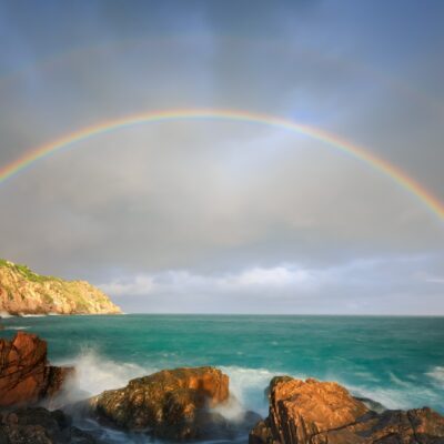A large rainbow stretches over the ocean near a cliff.